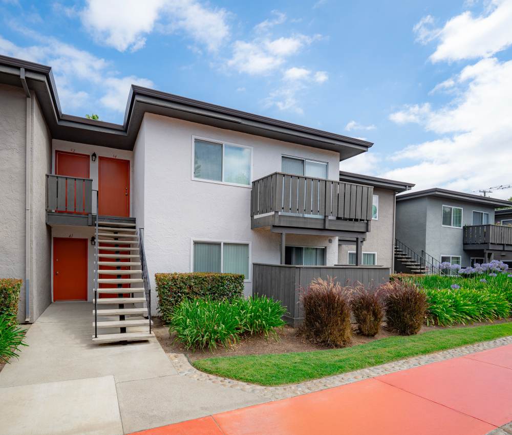 Exterior view of a two-story apartment building with balconies, colorful entrance doors, and well-maintained landscaping, set against a bright blue sky at Oliva in San Jose, California.
