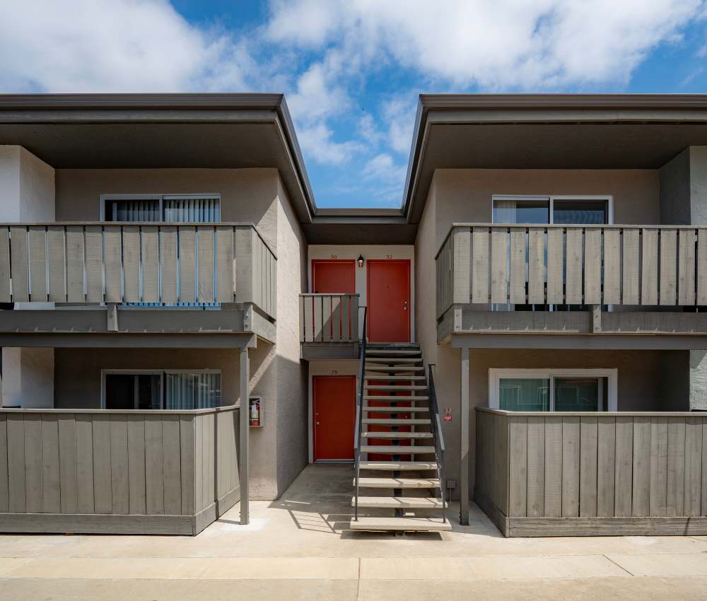 Front view of a two-story apartment building showcasing a staircase leading to bright orange doors, with balconies above and a clean pathway, set against a blue sky with scattered clouds at Oliva in San Jose, California.