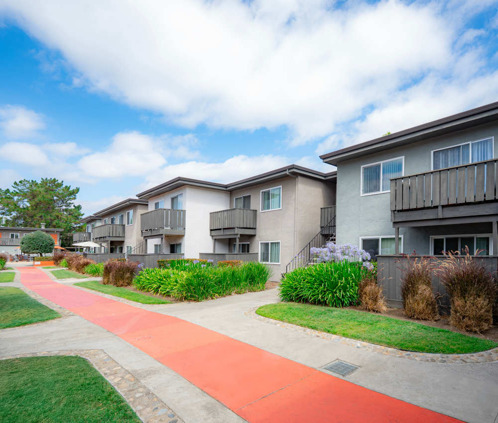 View of a residential complex with two-story apartment buildings featuring balconies, surrounded by landscaped pathways and vibrant greenery, under a blue sky with fluffy clouds at Oliva in San Jose, California.