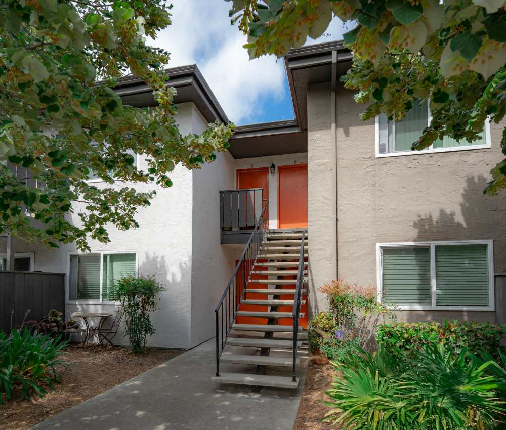 Close-up view of a two-story apartment entrance featuring a staircase, bright orange doors, and lush landscaping with greenery, framed by trees under a partly cloudy sky at Oliva in San Jose, California.