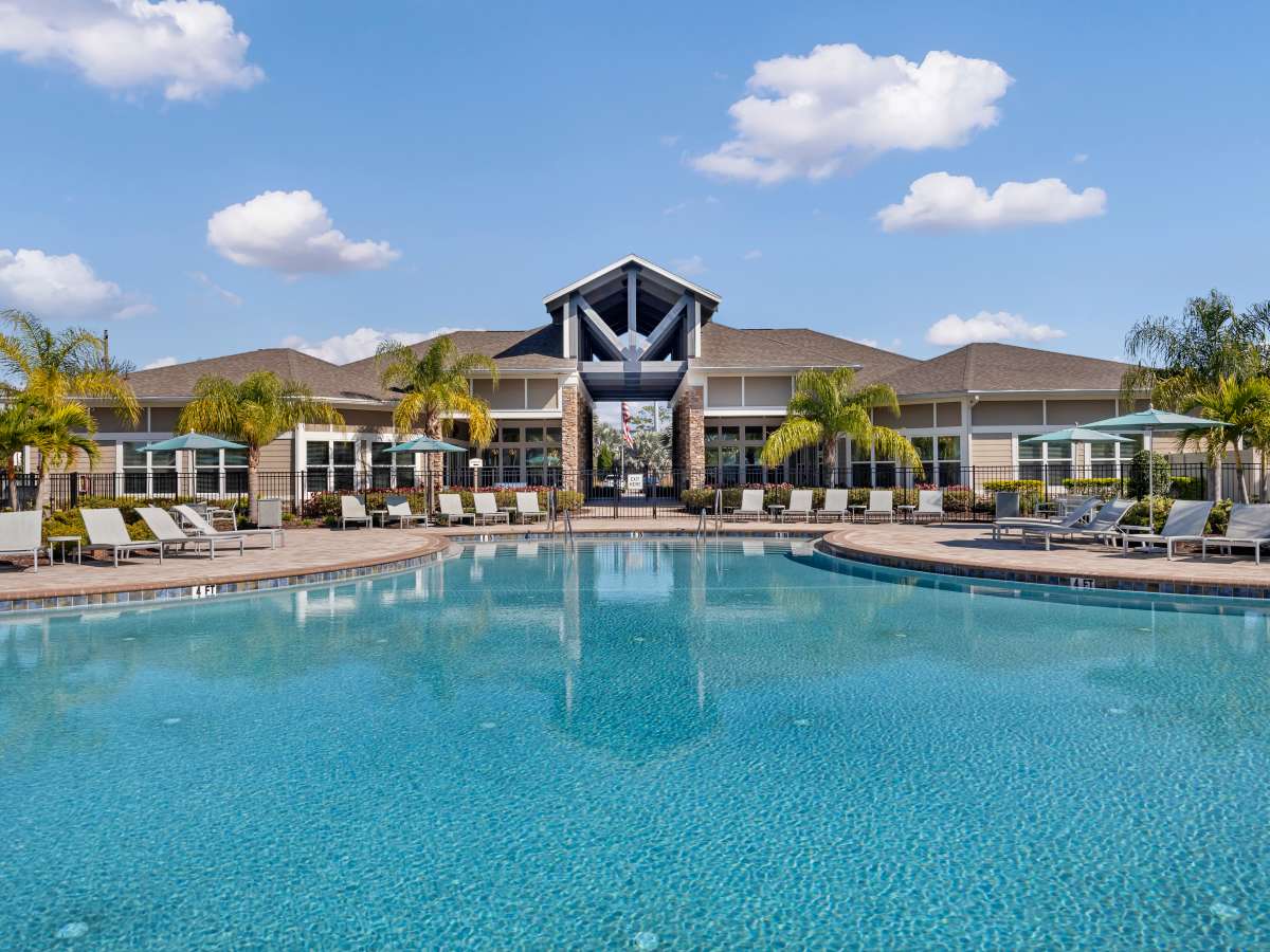 Resort-style pool surrounded by lounge chairs and palm trees with a clubhouse in the background at The Carlton of West Melbourne in West Melbourne, Florida