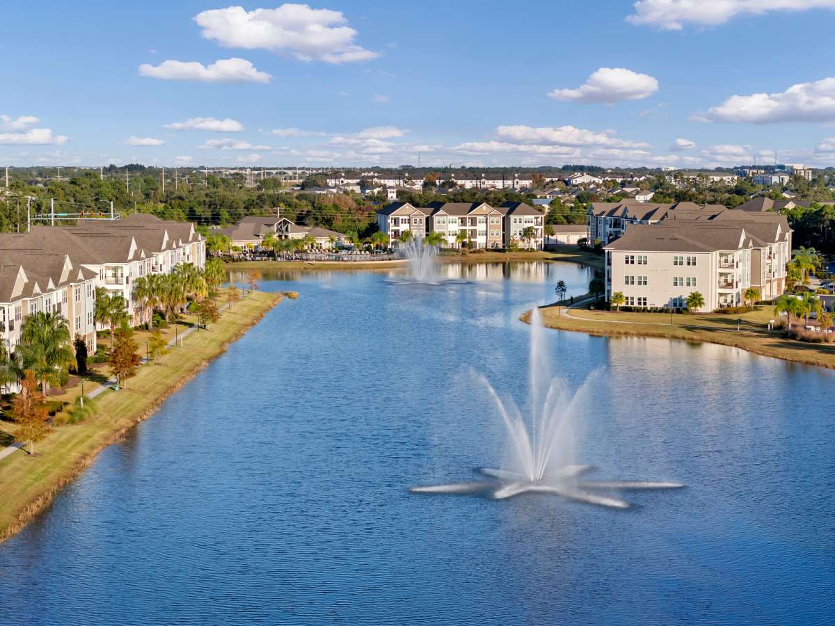 Lakeside view of apartment community with fountains and palm trees under a blue sky at The Carlton of West Melbourne in West Melbourne, Florida