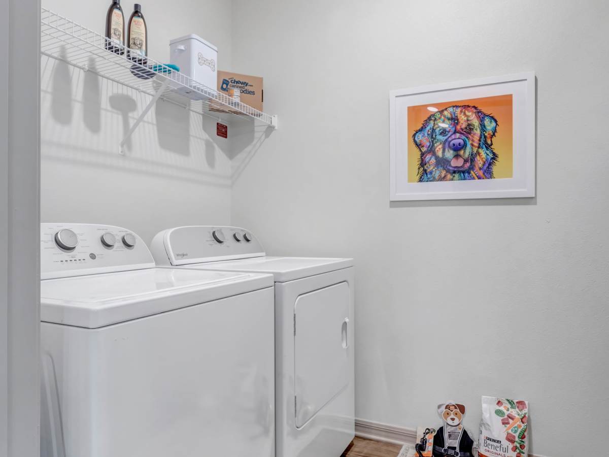 Laundry room with white appliances, pet-themed decor, and storage shelf above at The Carlton of West Melbourne in West Melbourne, Florida