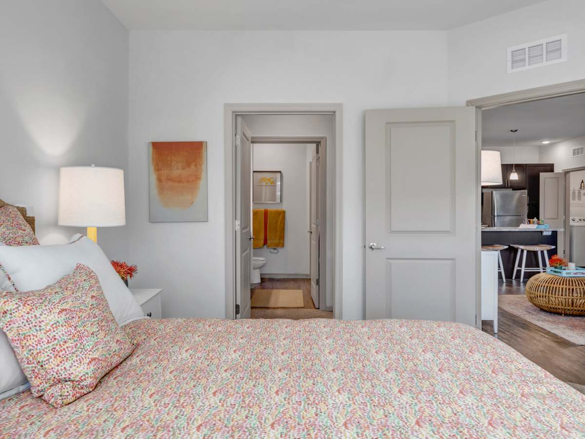 Cozy bedroom with colorful bedding, lamp, and open doorway to kitchen and bathroom at The Carlton of West Melbourne in West Melbourne, Florida