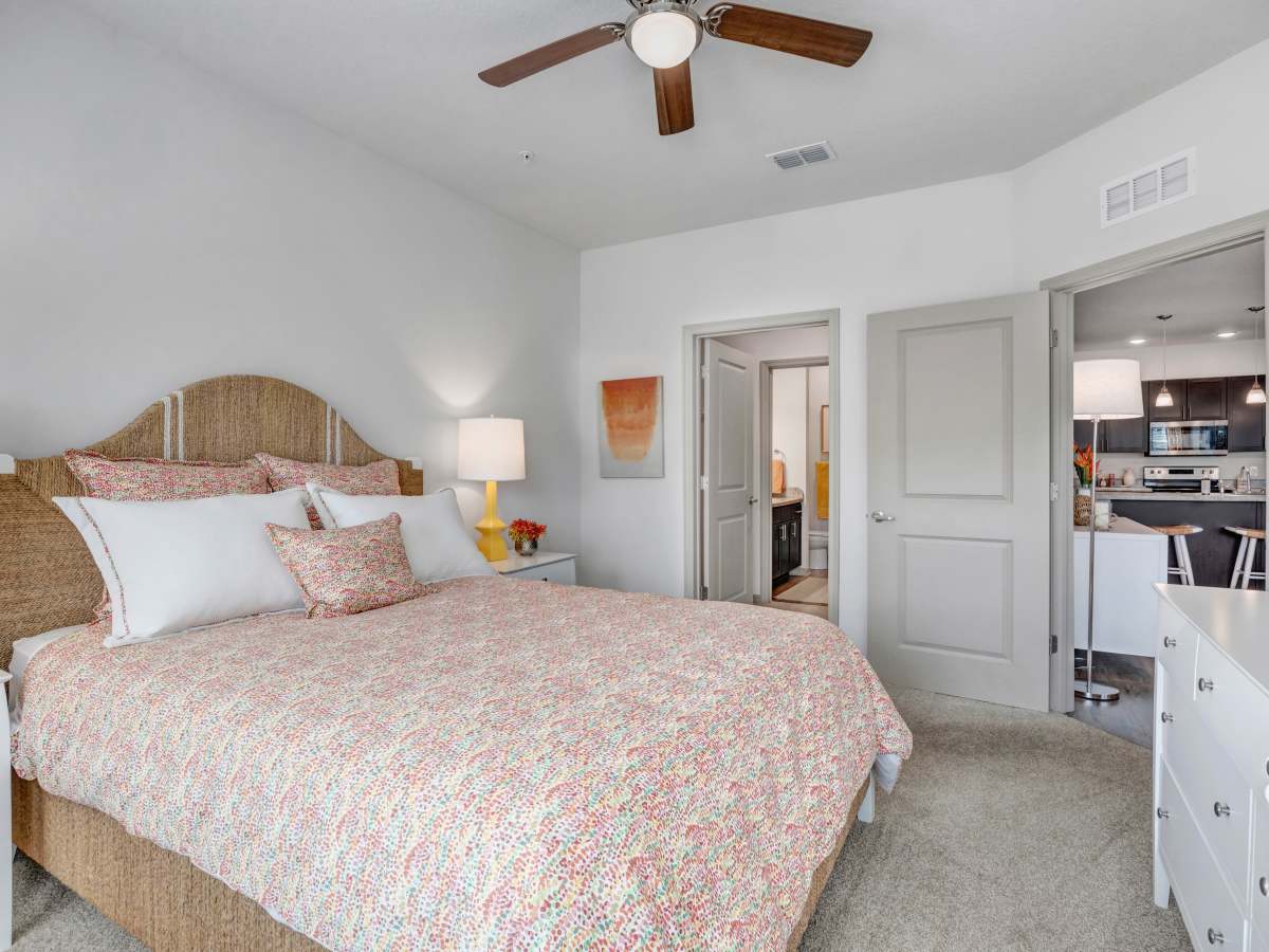 Spacious bedroom with a king-size bed, ceiling fan, and open layout to kitchen area at The Carlton of West Melbourne in West Melbourne, Florida