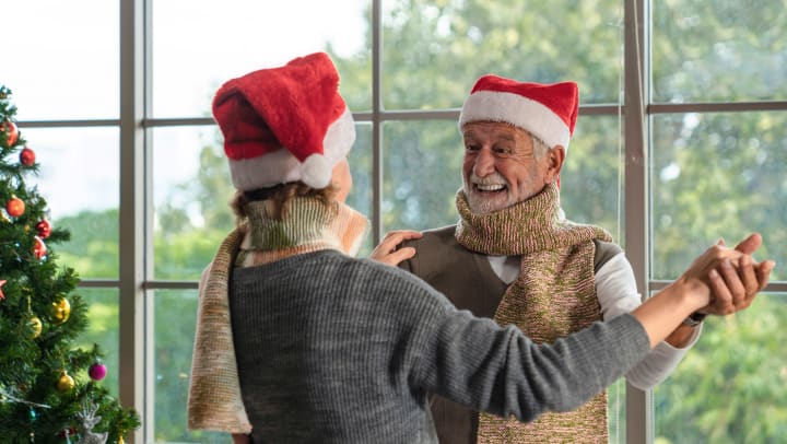 A senior man and woman dancing with Santa hats on with a large window and a Christmas tree in the background.