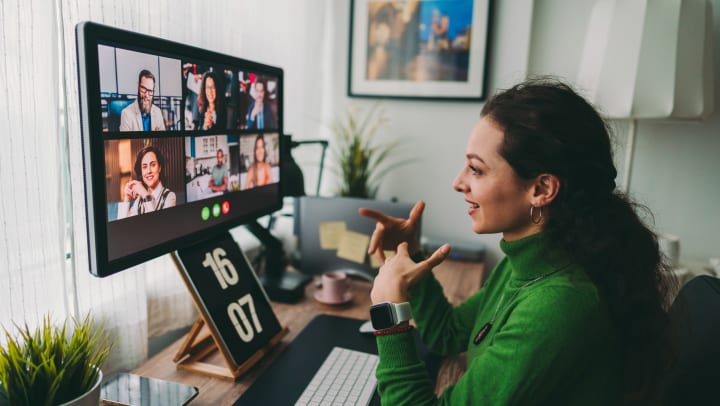 A woman sitting at a desk is looking at a computer where six others who are on a Zoom call with her.