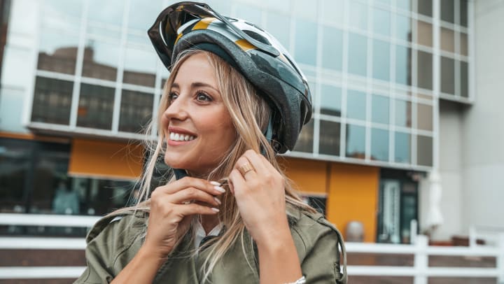 A woman is smiling and putting on a bike helmet with a building in the background