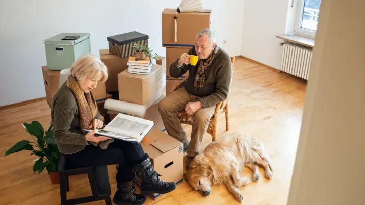 Two older adults sitting in chairs near stacks of moving boxes. A golden retriever is lying on the floor next to them.