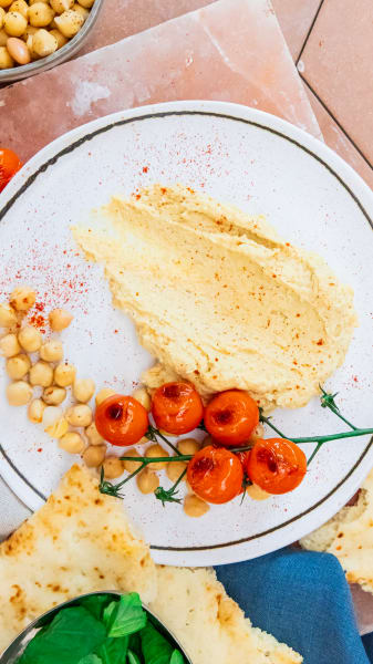 Spoon swoosh of hummus on a white plate garnished with garbanzo beans, a charred vine of cherry tomatoes, pita bread and a bowl of basil leaves