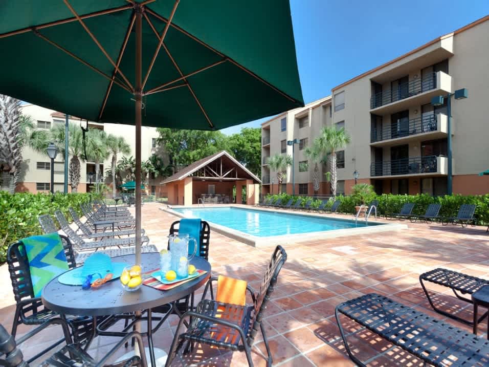 Sparkling swimming pool surrounded by lounge seating and patio tables with umbrellas at New Barn Apartments in Miami Lakes, Florida