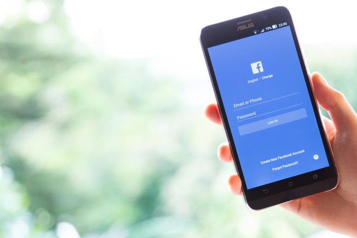 Photo of Asus smartphone with facebook app login screen being held by a hand against a blurred green vegetation background