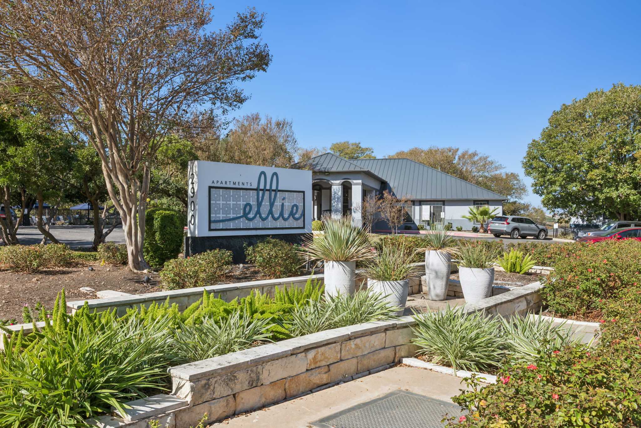 Entrance to Ellie Apartments featuring a large sign with stylized “ellie” text, the word “APARTMENTS” above it, and the address number “12900” on the side; surrounded by landscaped greenery, white planters, and a modern building with a dark metal roof in the background  at Ellie Apartments in Austin, Texas