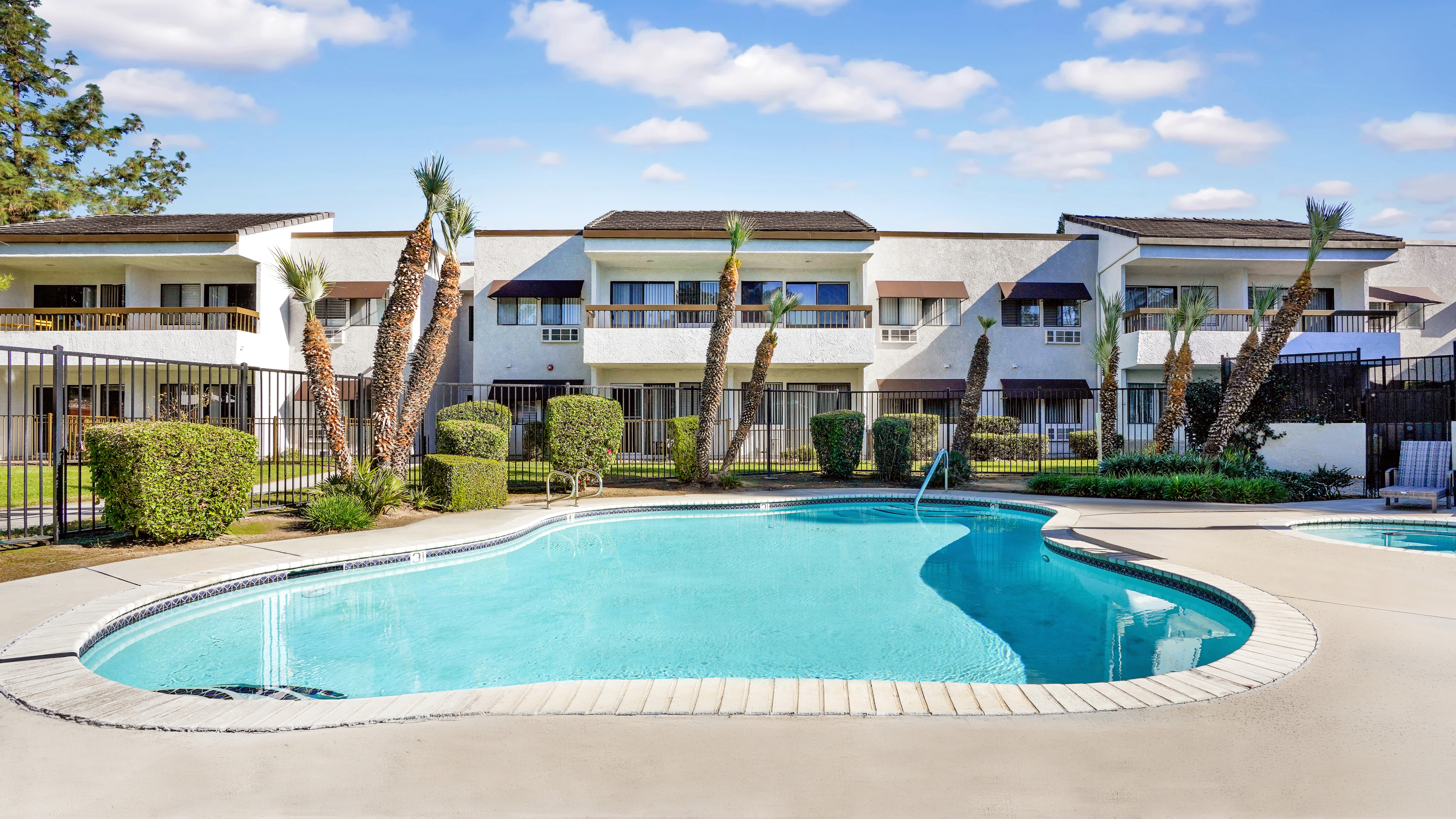 Outdoor swimming pool with curved edges surrounded by white decking, landscaped bushes, and multi‑unit residential buildings in the background at Citrus Place in Riverside, California