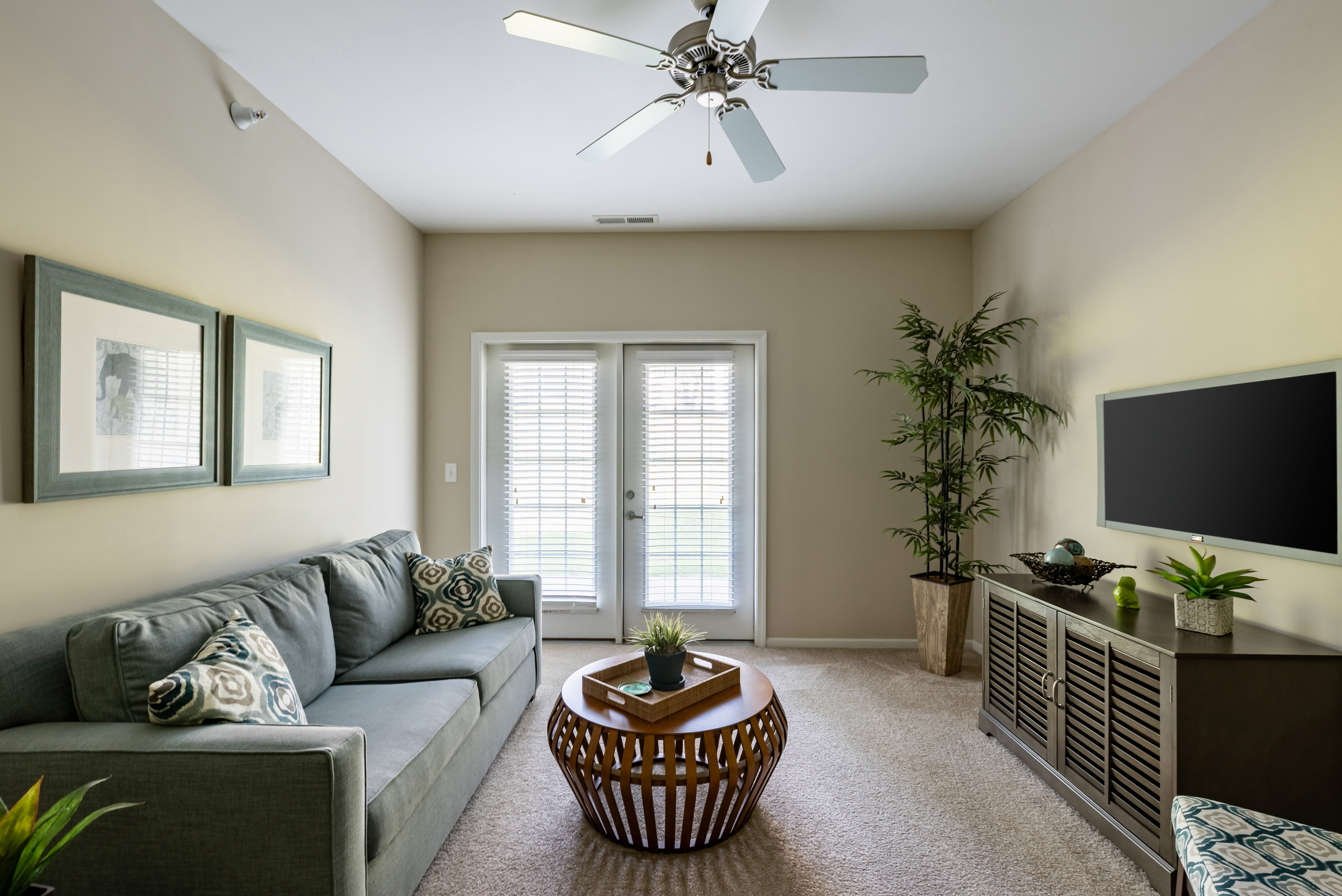 Living room with gray sofa, patterned pillows, coffee table, ceiling fan, and double glass doors leading outside at Oak Grove Crossing Apartments in Newburgh, Indiana.