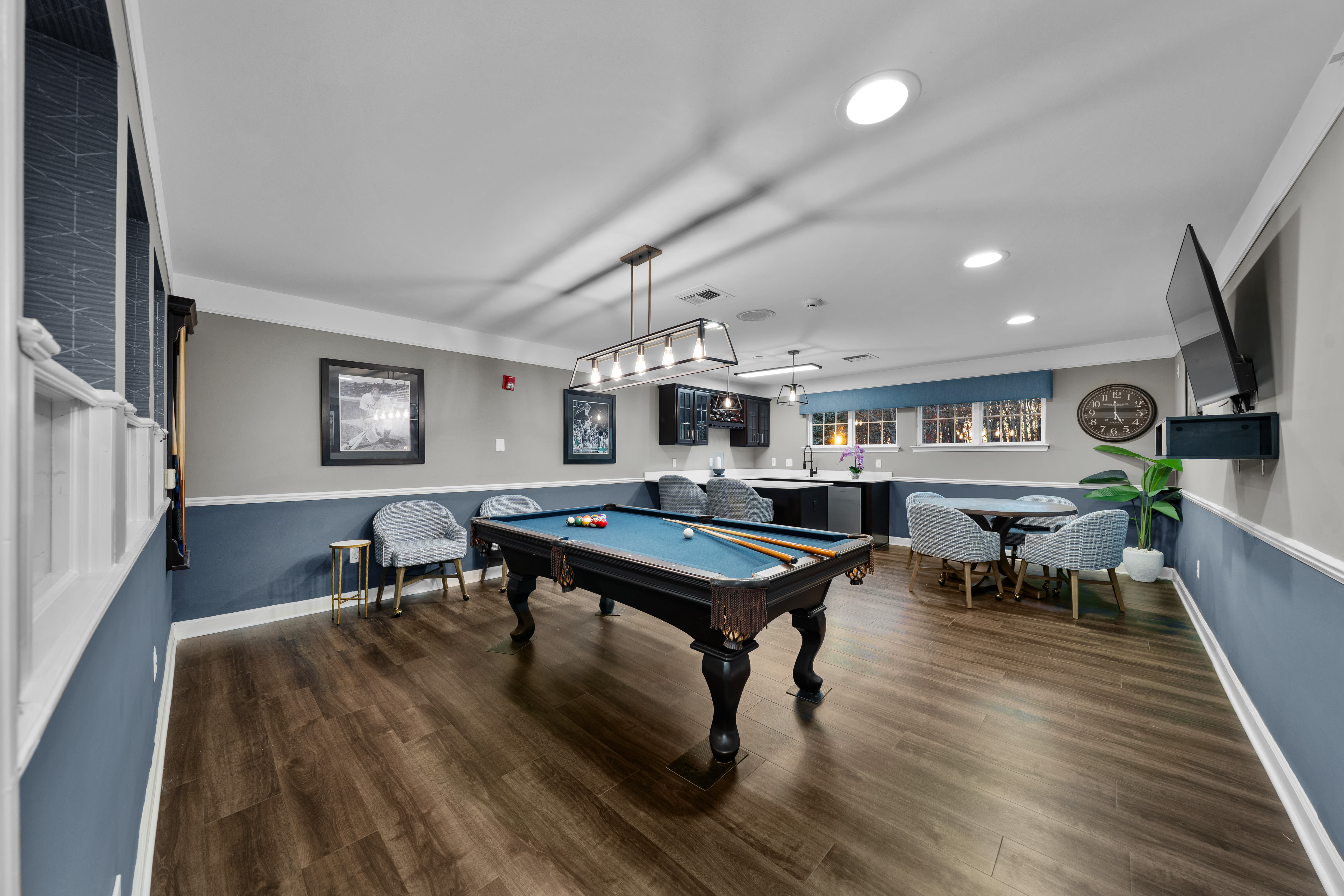 Game room featuring a pool table with arranged billiard balls, overhead rectangular light fixture, wood‑look flooring, upholstered chairs, wall art, mounted television, and a seating area with tables near the windows at Waltonwood Cherry Hill in Canton, Michigan