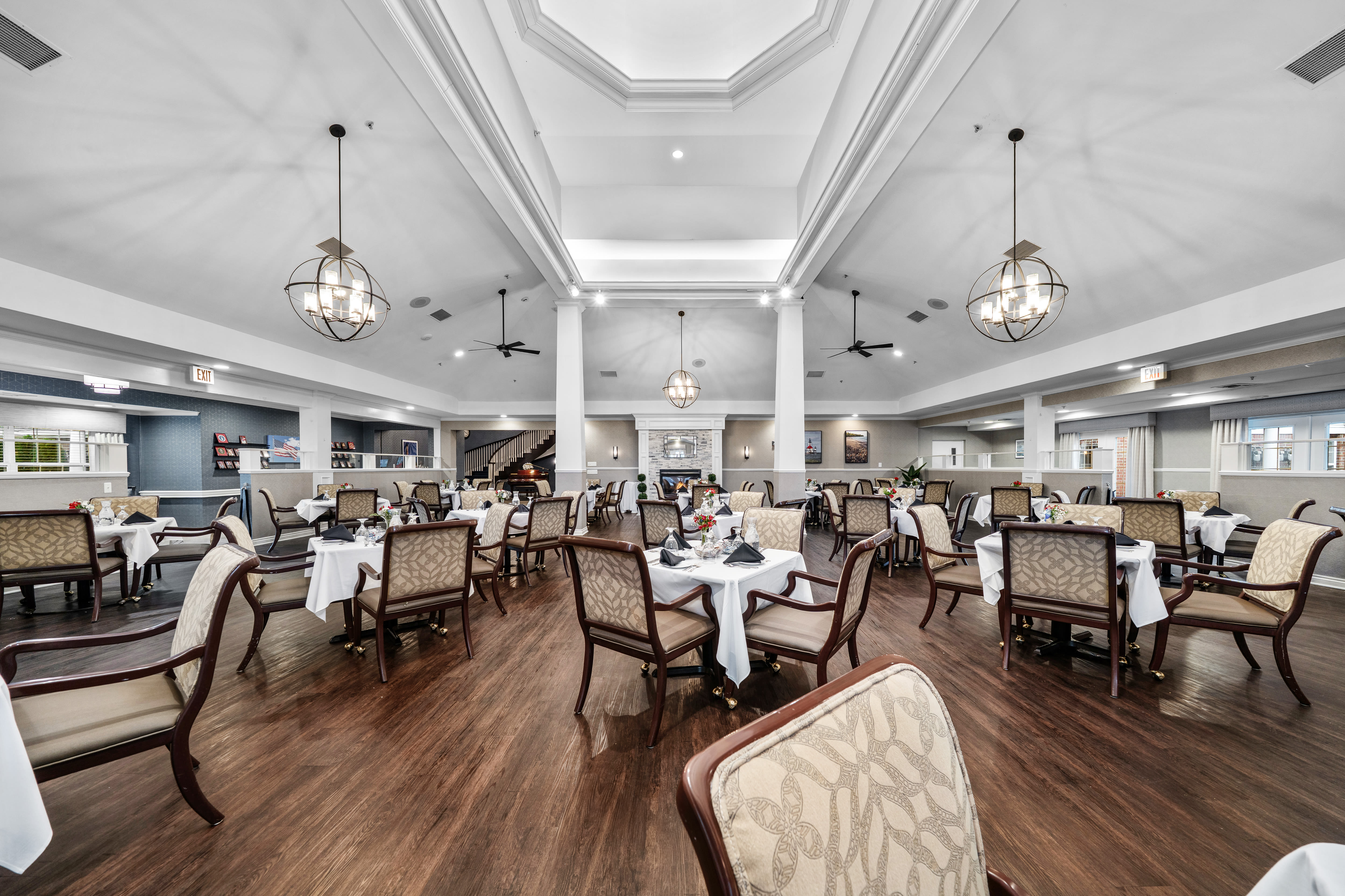 Spacious dining room with neatly arranged tables covered in white tablecloths, cushioned chairs, elegant pendant lighting, tall white columns, and hardwood flooring at Waltonwood Cherry Hill in Canton, Michigan