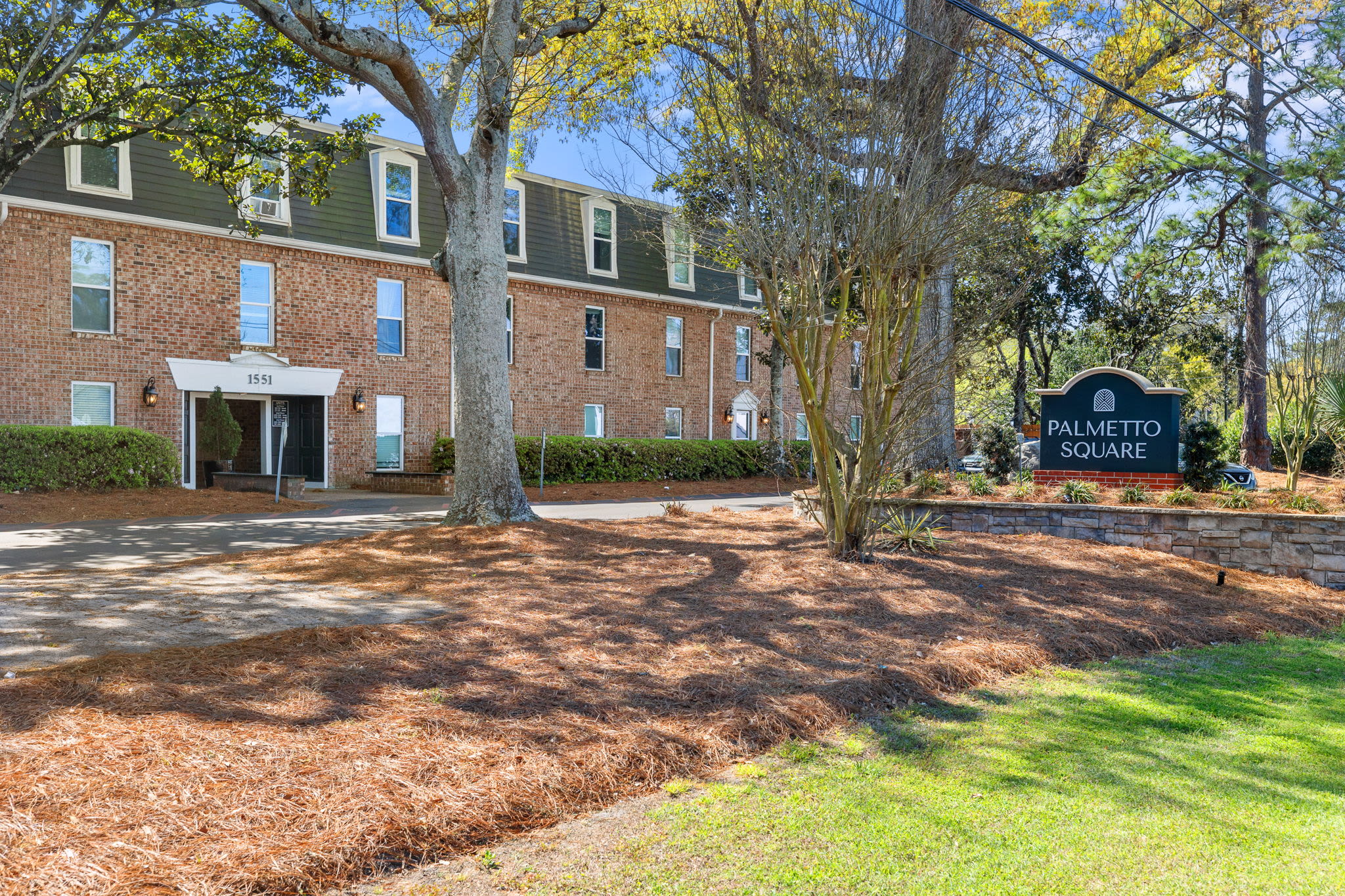 Apartment entrance with Palmetto Square sign, landscaped lawn, and brick residential building at Palmetto Square in Charleston, South Carolina