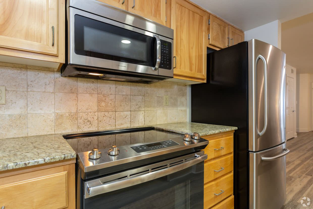 Kitchen with wooden cabinetry, granite countertops, built‑in microwave, electric cooktop, stainless steel refrigerator, tiled backsplash, and wood‑look flooring extending into a hallway at Oakshade Commons in Davis, California.