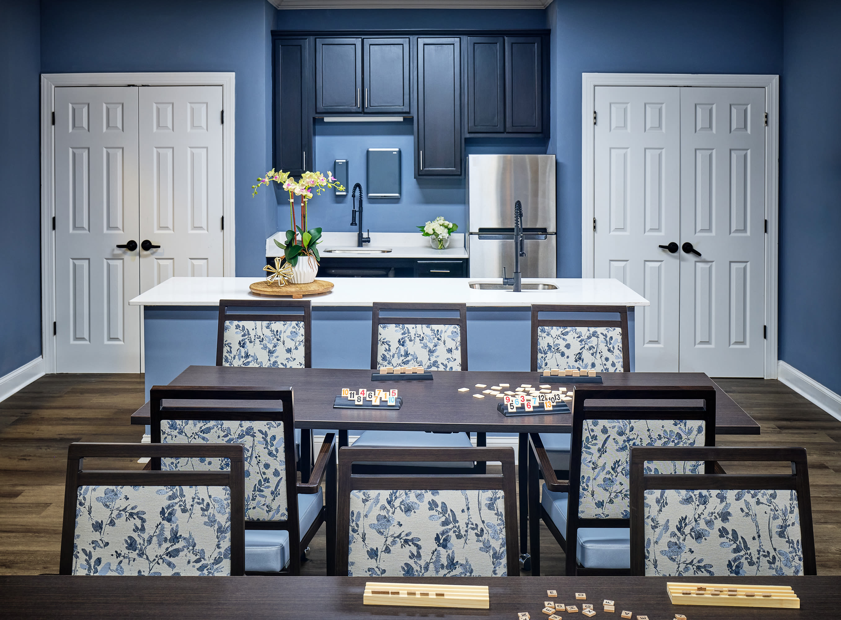Community clubhouse dining area with blue accent walls, communal dining table, kitchen island seating, stainless steel appliances, and modern cabinetry at Waltonwood Cotswold in Charlotte, North Carolina