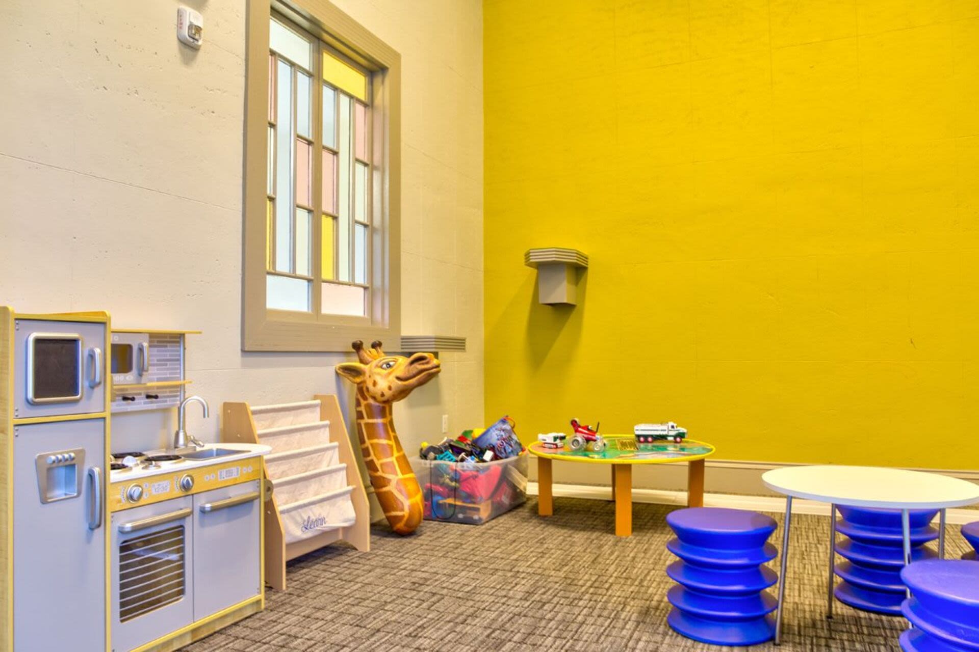 A playroom with a kitchen set, a giraffe toy, a small table, and toy cars at Bethany Apartments in Hanover, Massachusetts
