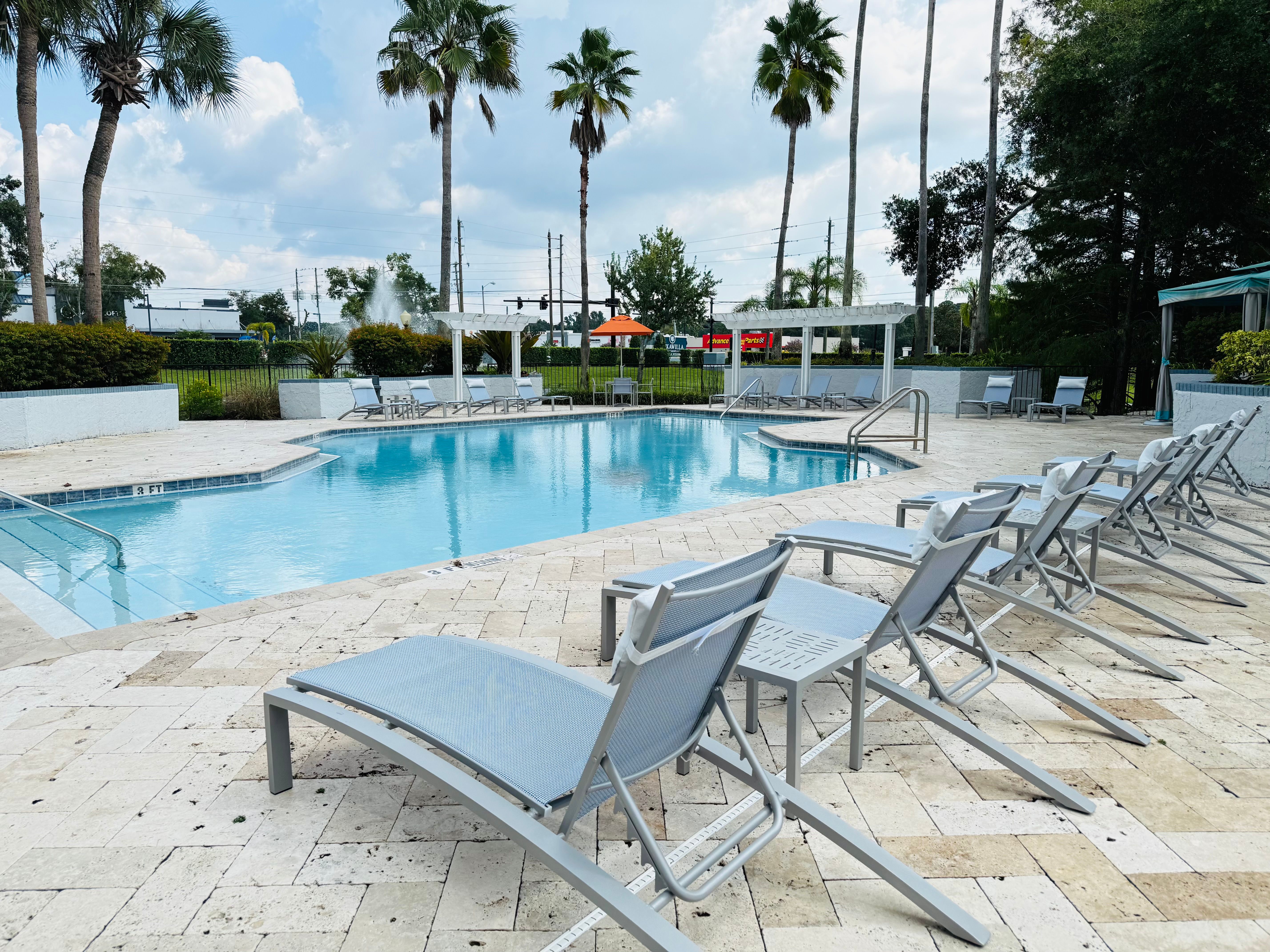 Poolside lounge chairs beside a clear outdoor swimming pool with palm trees in the background at Tuskawilla at Winter Springs in Winter Springs, Florida