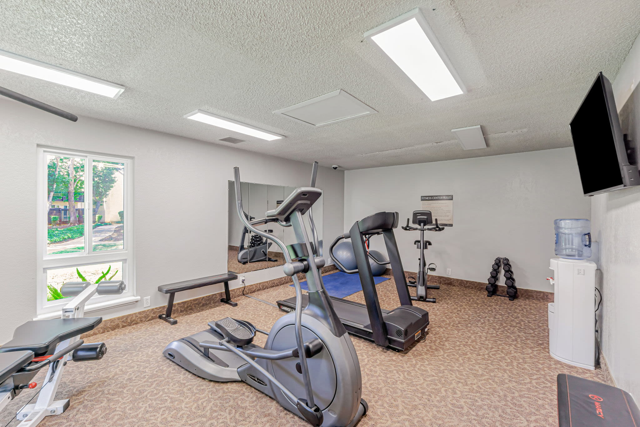 Small fitness room with cardio machines, weights, and a bench, shown in a bright, clean space at Clayton Gardens in Concord, California