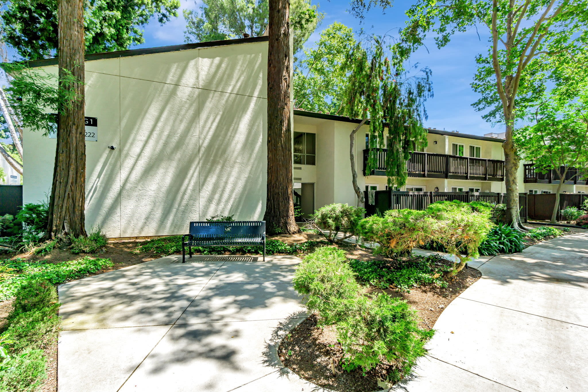 Shaded walkway with benches, tall trees, and residential buildings surrounded by greenery at Clayton Gardens in Concord, California