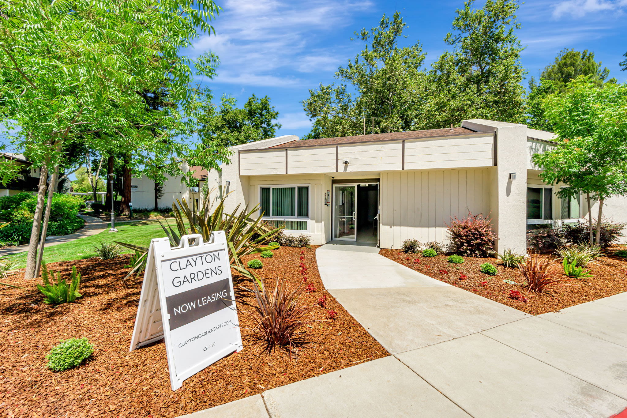 Leasing office entrance with a now‑leasing sign, landscaped plants, and walkway at Clayton Gardens in Concord, California