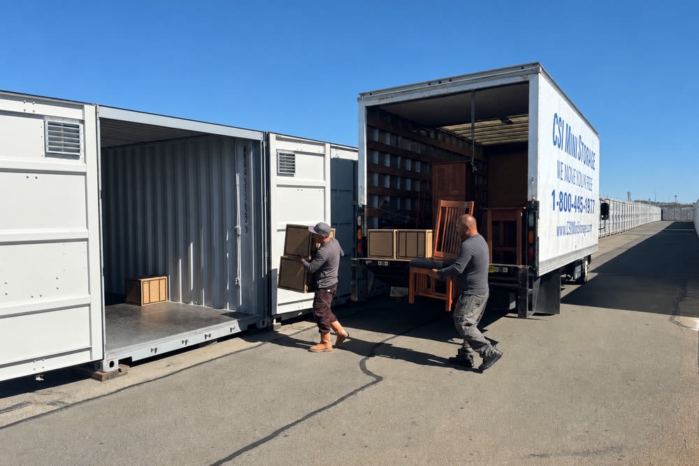 Two movers unloading furniture from a CSI Mini Storage truck into a drive-up storage unit in Alameda.