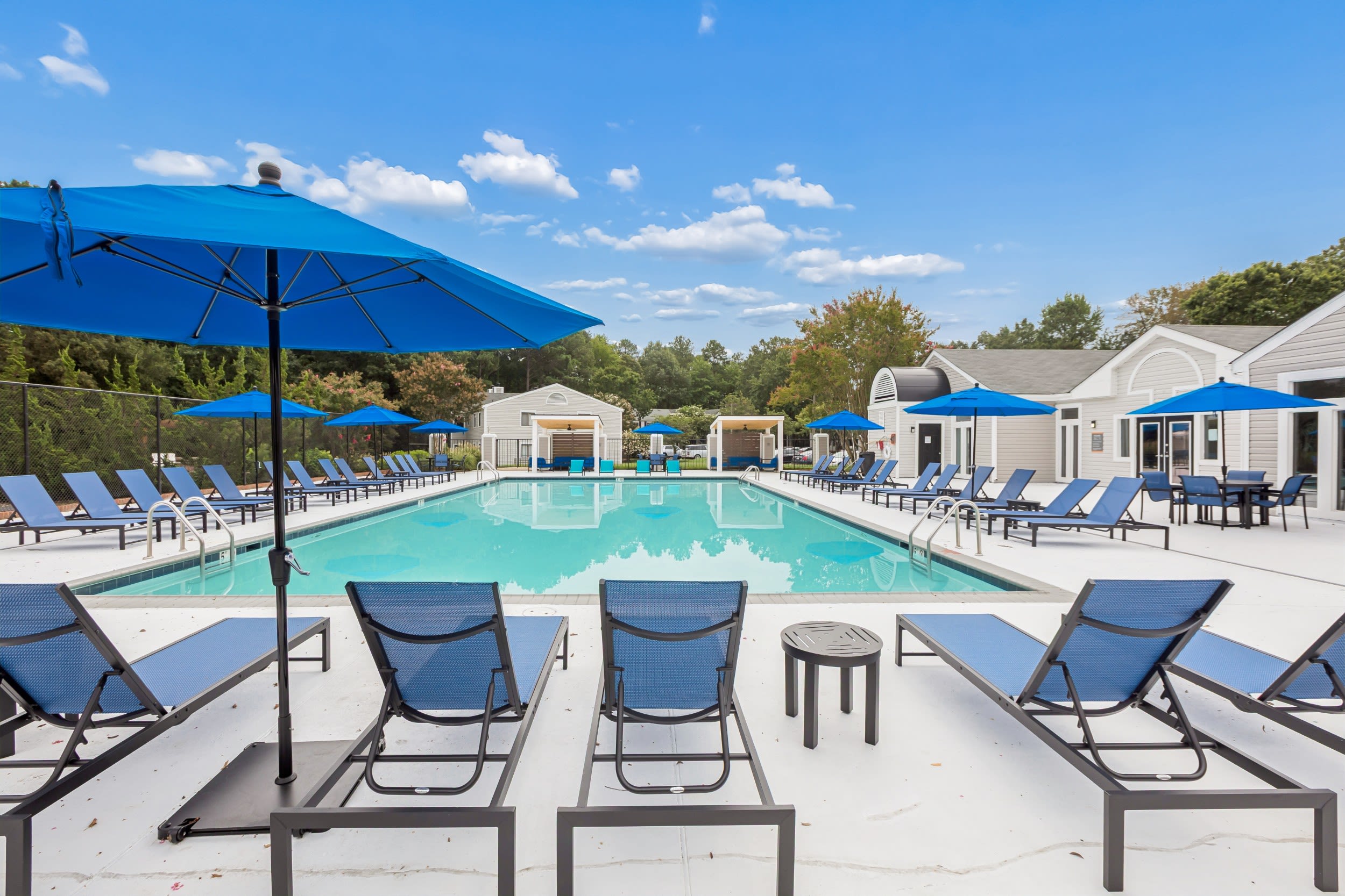 Reserve South - Resort-Style Swimming Pool With Sundeck, Lounge Chairs, Umbrellas, and Clubhouse Buildings Surrounded By Trees Under Blue Sky.