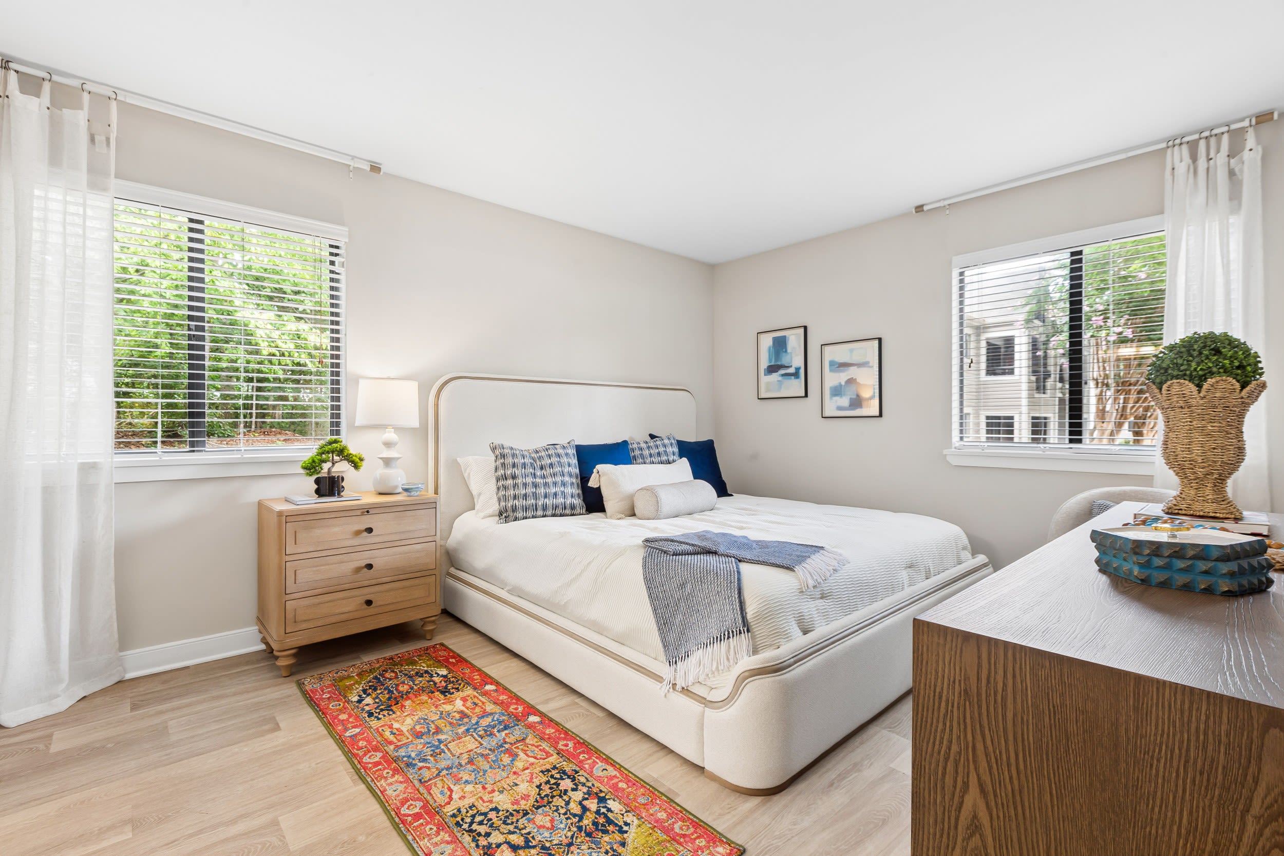 Reserve South - Bright Bedroom With Upholstered Bed, Nightstands, Table Lamps, Large Window Blinds, and hardwood-style flooring.