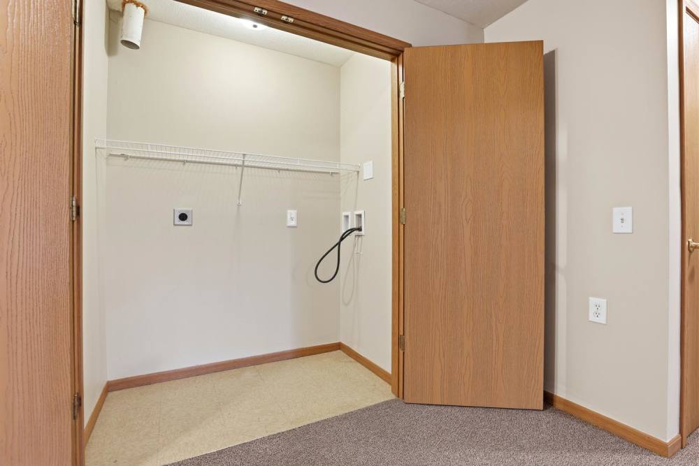 Empty laundry room with shelving and utility connections, beige walls and carpeting at Norwood Greene Senior Living in Marietta, Ohio