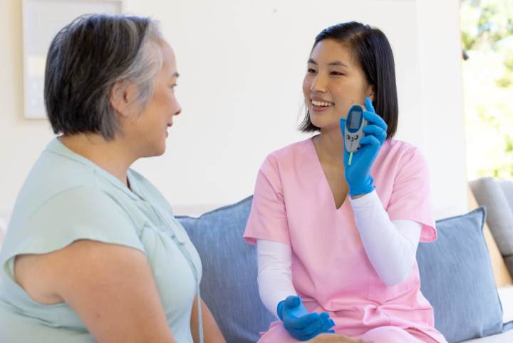 Senior diabetes care: A nurse in pink scrubs assisting an elderly woman with glucose monitoring in a home setting.