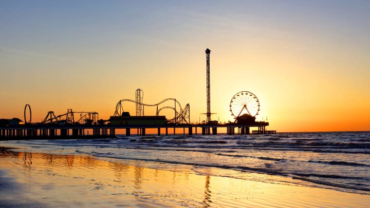 A ferris wheel and other rides are silhouetted by the sunset, with waves in the foreground.