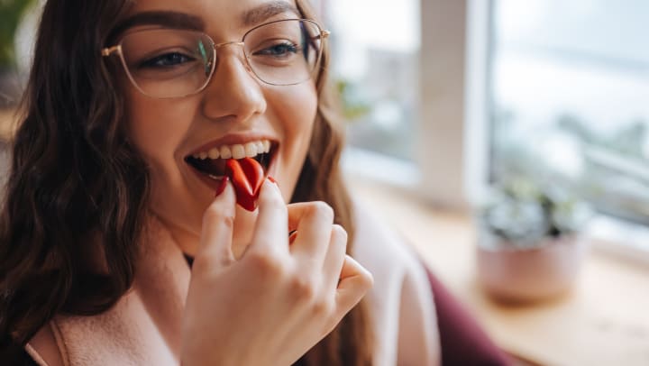 A smiling young woman with eyeglasses putting a piece of red candy in her mouth.