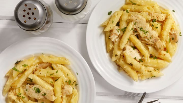 Two ceramic plates of chicken alfredo with penne pasta on a white wooden table.