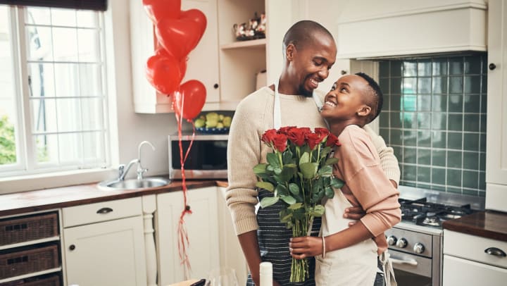 A young couple wearing aprons hug in the kitchen as she holds a bouquet of roses, with two glasses of wine on the counter among their ingredients, and red heart-shaped balloons in the background.
