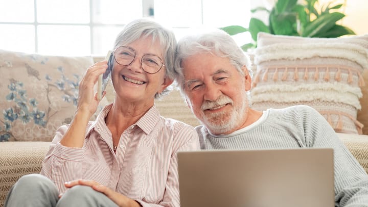  A smiling older couple in their living room, the man looking at a laptop computer and the woman on a mobile phone.