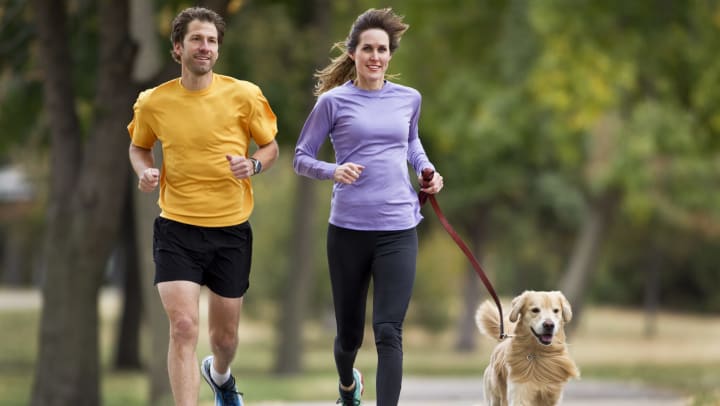 A smiling man and woman with a leashed golden retriever are jogging on a paved trail. 