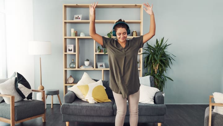 A happy young woman using headphones and dancing in the living room at home.