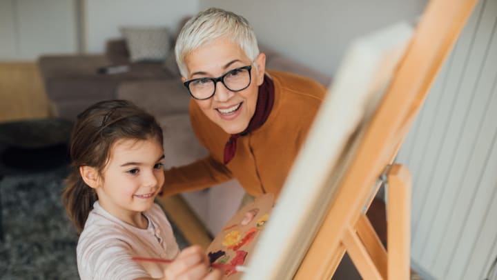A smiling older woman holding an artist’s palette and standing next to a young girl who is holding a paintbrush and looking at a canvas