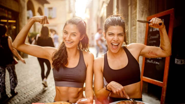Two sporty women eating pizza. They are flexing their muscles while looking at the camera.