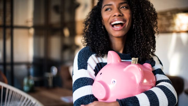 Woman smiling while hugging a pink piggy bank with a bill sticking out of the top. 