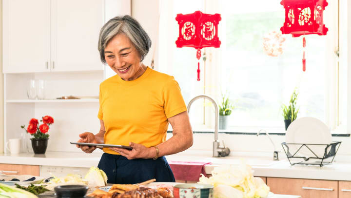 Woman holding an ipad and looking at a countertop full of food with red paper lanterns hung in a window behind her. 