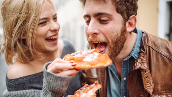A laughing young man and woman feed each other slices of pizza. 