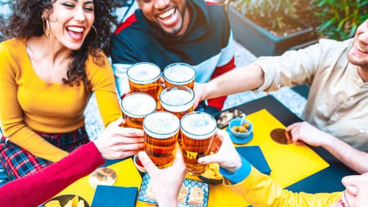 Group of men and women smiling while holding full pints full of beer up together over a yellow tabletop. 