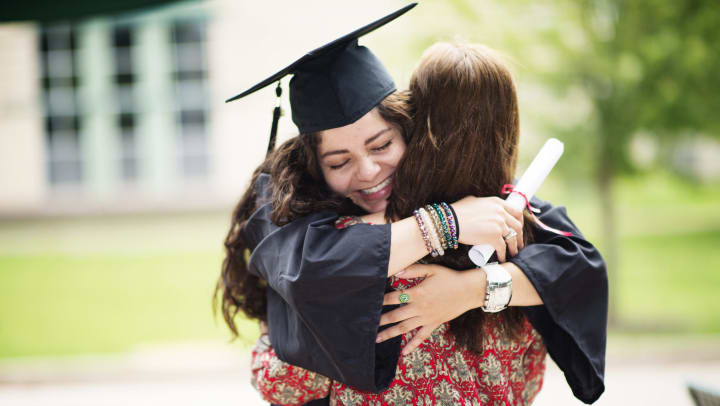 Two females hugging. One has a cap and gown on and is holding a diploma. 