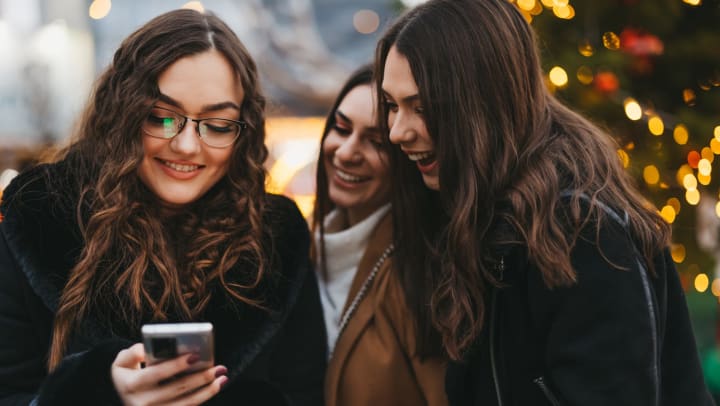 Young women looking at a smart phone and smiling with lights out of focus behind them.