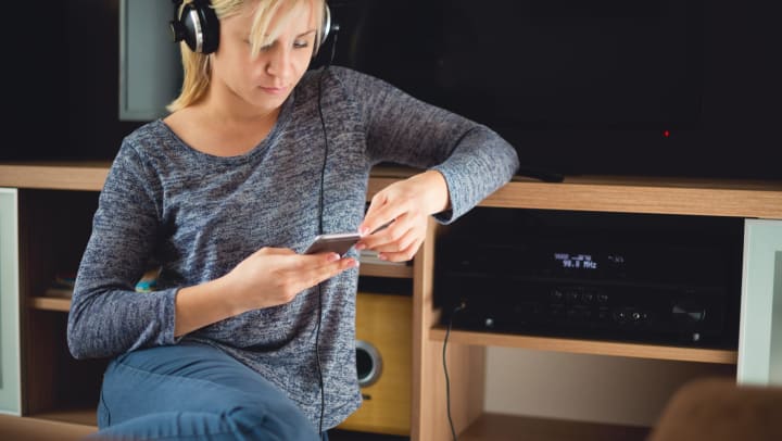A woman sitting on the floor next to an entertainment center. She has headphones on while looking at her phone. There’s a TV next to her and it is off.